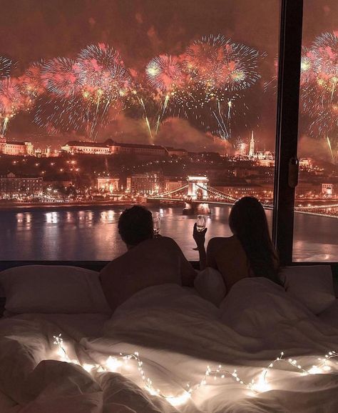 Couple in bed watching colorful fireworks over illuminated city skyline through large window