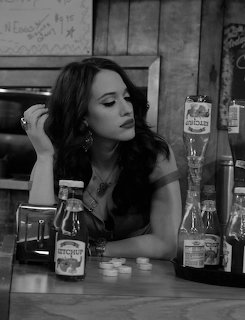 Black-and-white photo of a woman seated at a diner counter with bottles on the table.