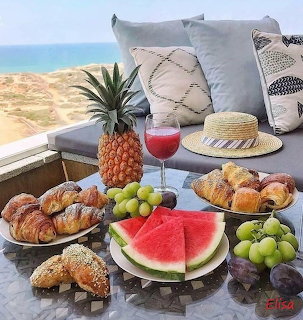 Pastries and fruit on a seaside table with a straw hat, cushions, and ocean view