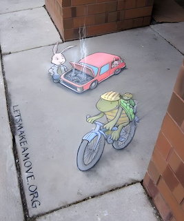 Chalk street art of a frog riding a bicycle and a white rabbit driving a pink car on a sidewalk