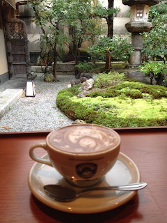 Coffee cup with latte art on a wooden ledge overlooking a traditional Japanese garden with stone lanterns and moss