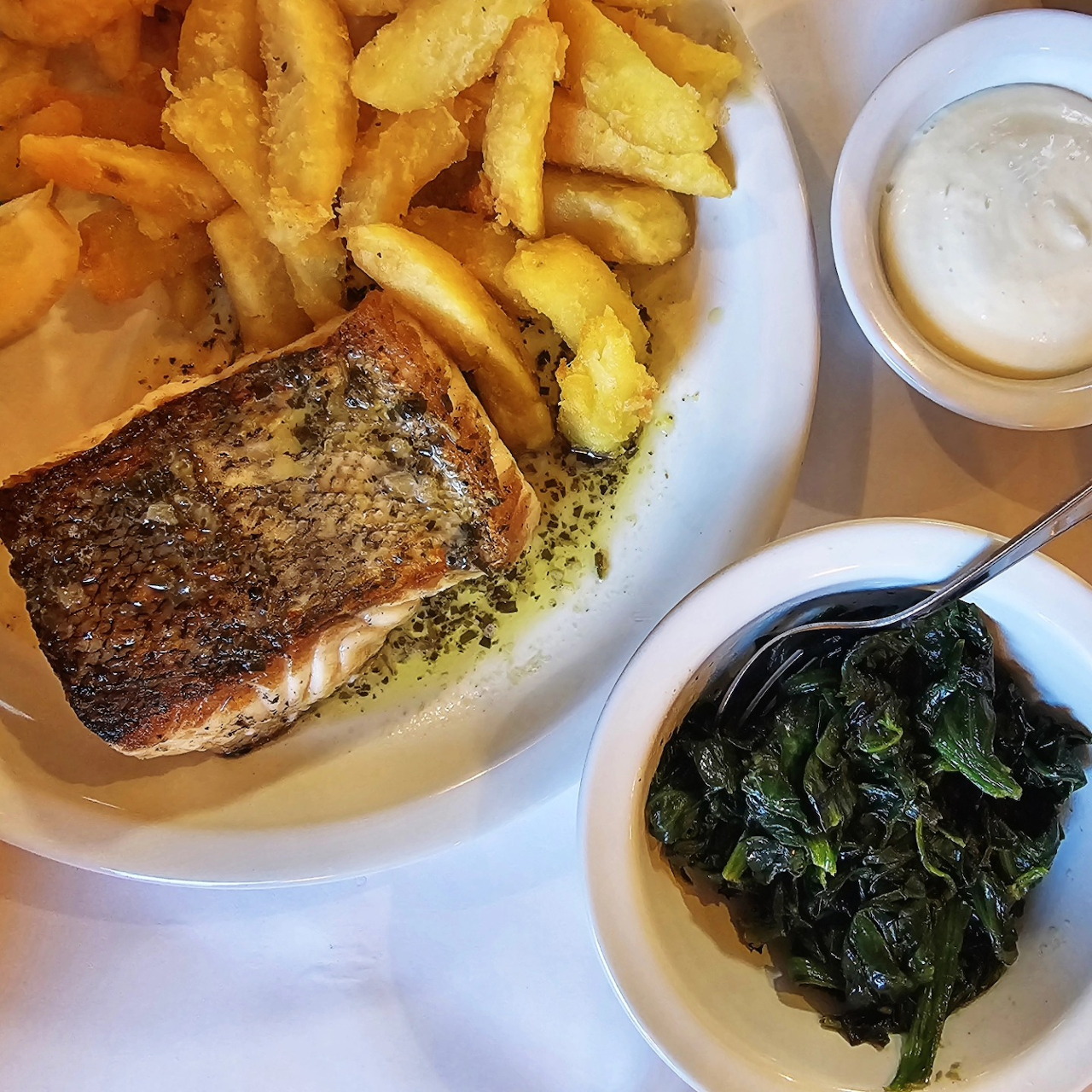 Seared salmon with French fries, creamed spinach, and a small cup of sauce on white plates
