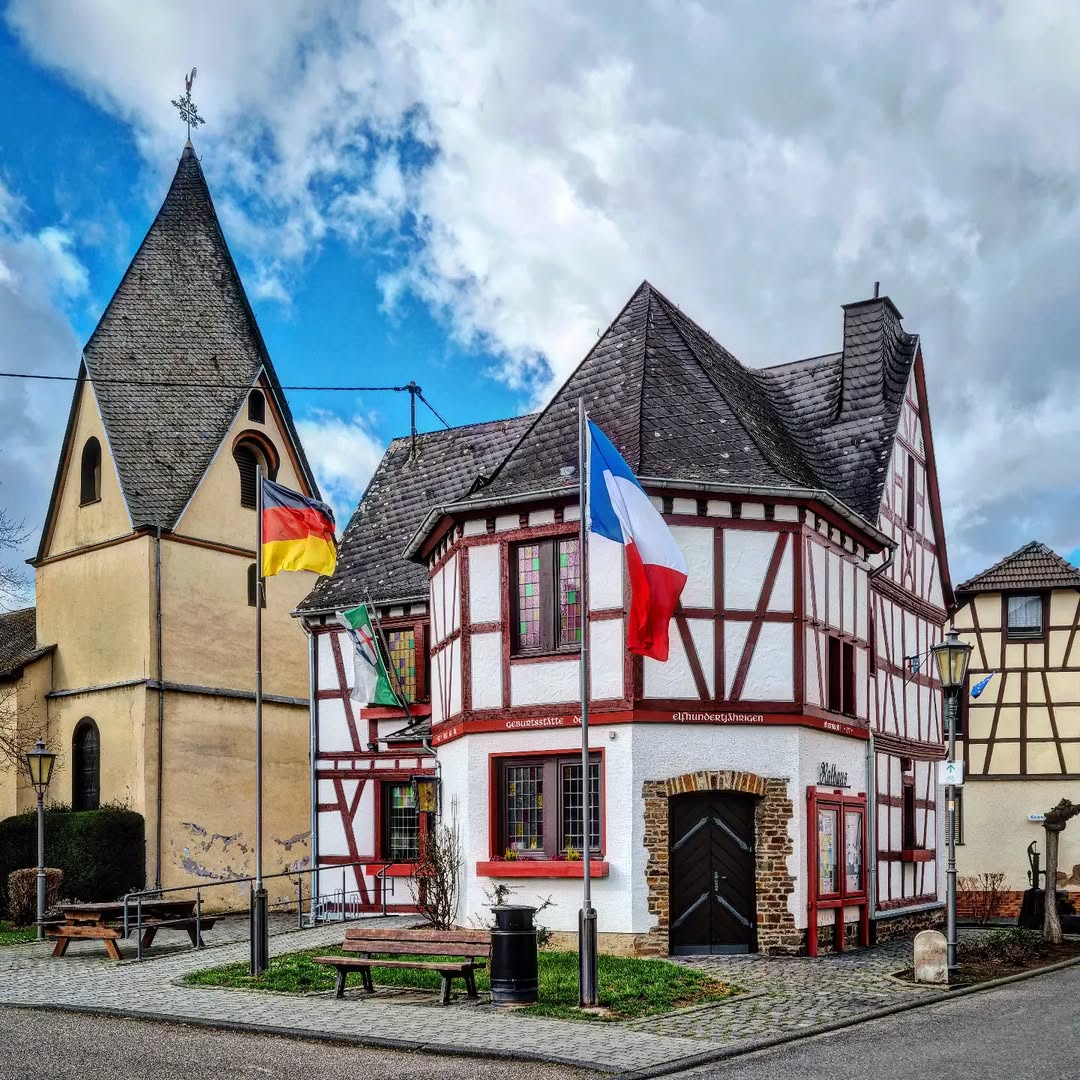 Traditional German village square with half-timbered houses, church tower, and national flags under cloudy blue sky