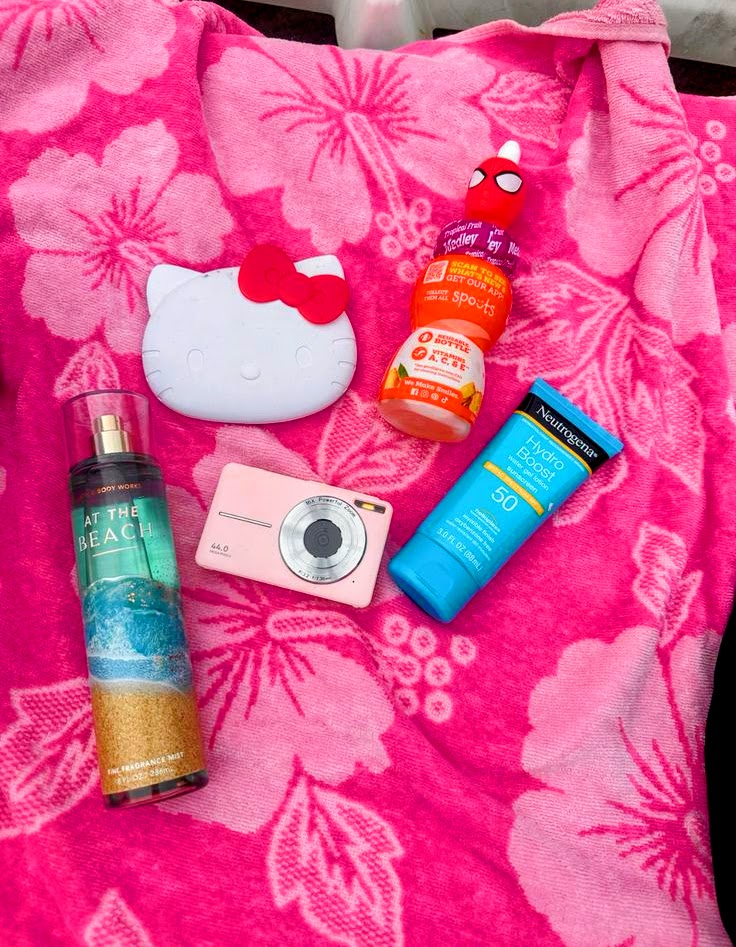 Pink beach towel with Hello Kitty case, compact camera, body mist spray, sunscreen tube, and lip balm flat lay