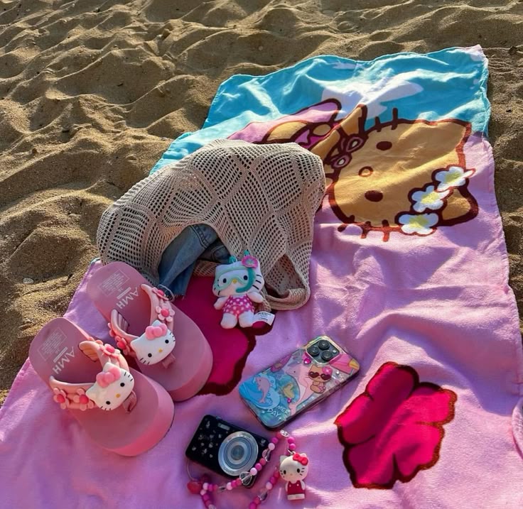 Hello Kitty themed beach items on sand with pink sandals, towel, phone case, camera, and small figures