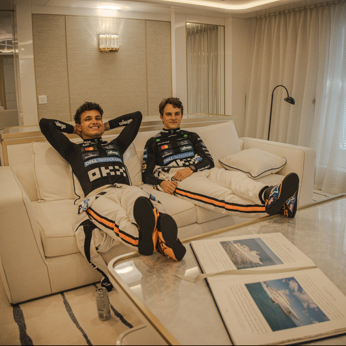Two McLaren F1 drivers in race suits lounging on a sofa in a modern living room