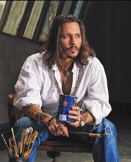 Man with long hair in white shirt holding paintbrushes and blue paint can in studio setting