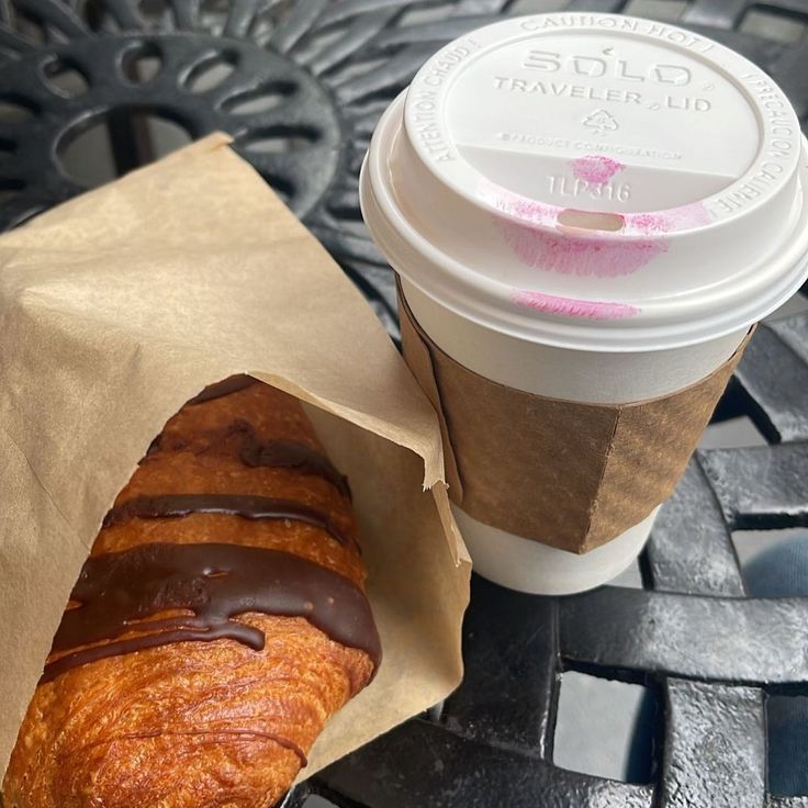 Takeaway coffee cup with lipstick marks beside a chocolate-drizzled croissant on a metal table
