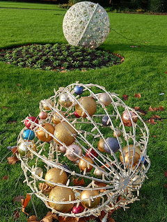 Large wire sphere filled with colorful Christmas baubles on grass, with a white wire ball decoration in the background.