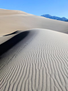 Large sand dune with wind-ripled texture and mountains in the background under blue sky
