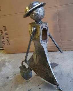 Welded metal sculpture of a humanoid figure wearing a hat and holding a padlock, made from scrap metal parts