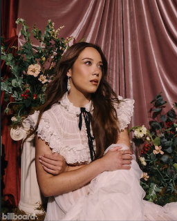 Woman in a white ruffled dress seated among flowers with pink satin drapes behind her.