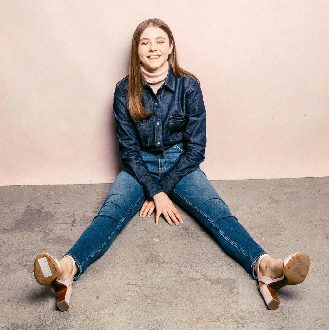Young woman in denim jacket and jeans sitting on gray floor with legs spread, smiling at camera