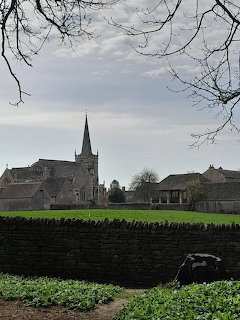 Village church with tall spire behind stone wall and hedge, bare trees frame cloudy sky