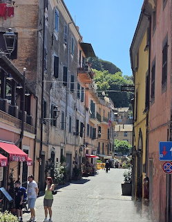 Narrow street in Nemi, Italy with colorful buildings, pedestrians, and a green hillside in the background