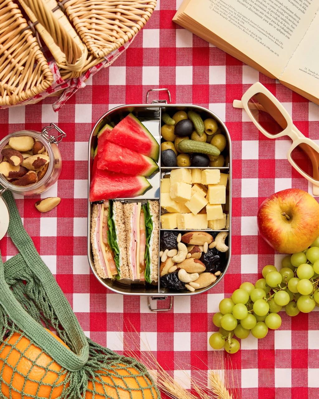 Overhead view of a picnic bento box with sandwiches, watermelon, cheese and nuts on a red checkered blanket with grapes and apple