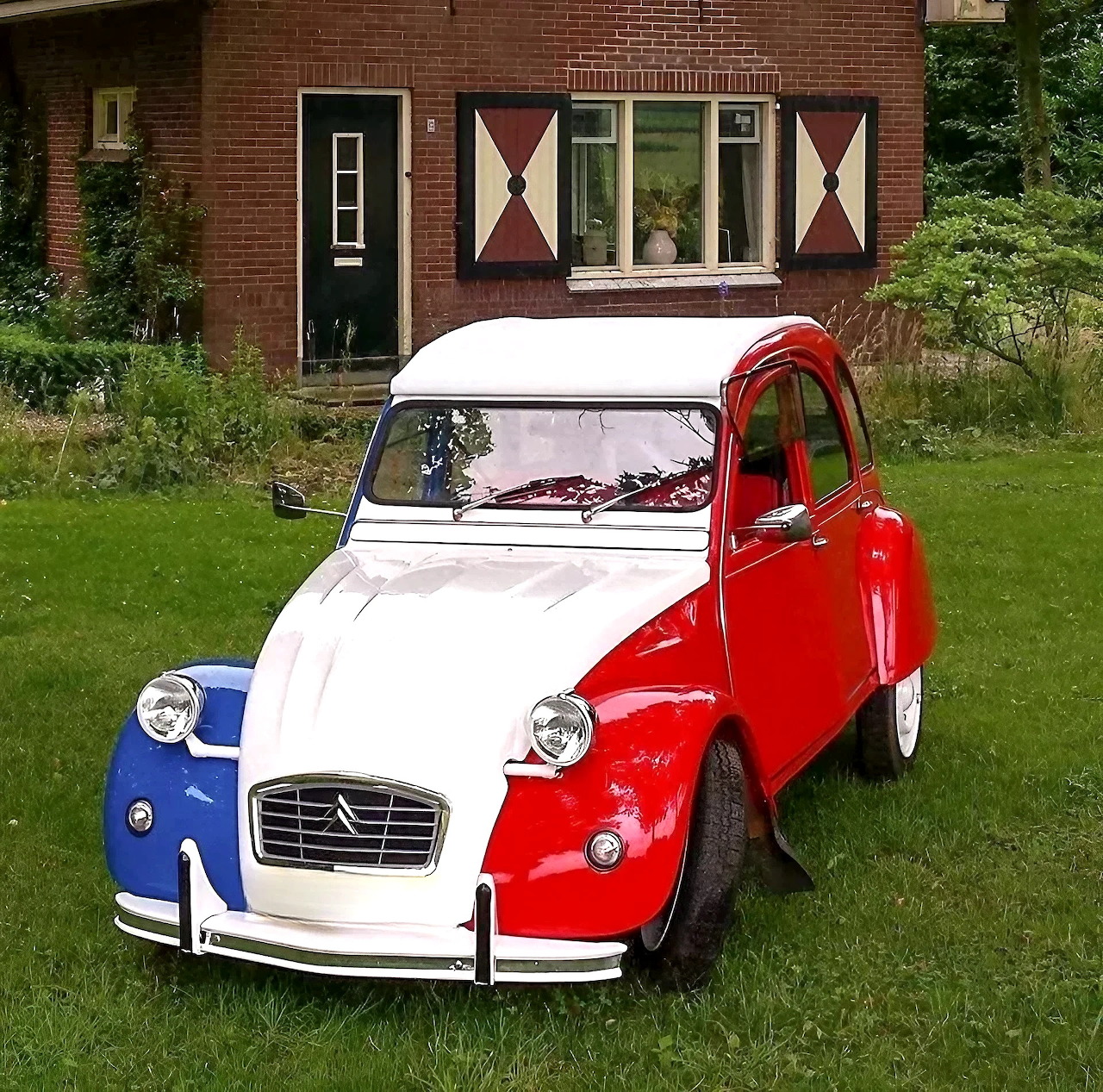Red, white and blue striped Citroën 2CV classic car parked on grass in front of brick house