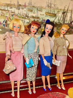 Four vintage Barbie dolls in colorful 1960s outfits standing in a row against a nautical harbor painting backdrop