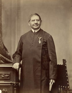 Black and white vintage portrait of man in dark clerical robes standing beside furniture