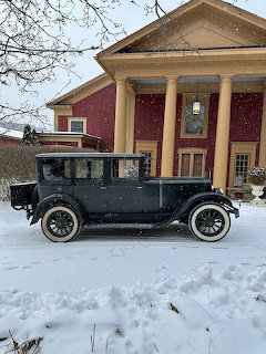 Black antique car parked in snow in front of a red and yellow mansion with columns