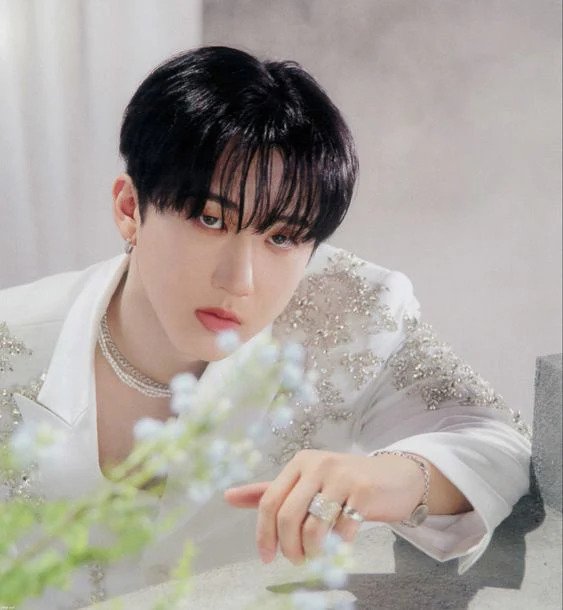 Young man with dark hair in white sparkly outfit posing near white flowers with silver jewelry