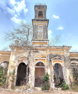 Abandoned old building with a clock tower, arched entrances, and sparse vegetation