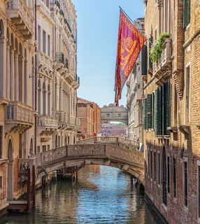 Narrow Venice canal with stone bridge, old buildings, and a red Venetian flag on a sunny day