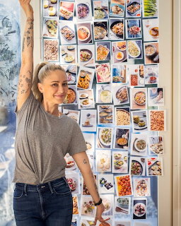 Woman chef in gray shirt standing next to wall grid of food photos in kitchen