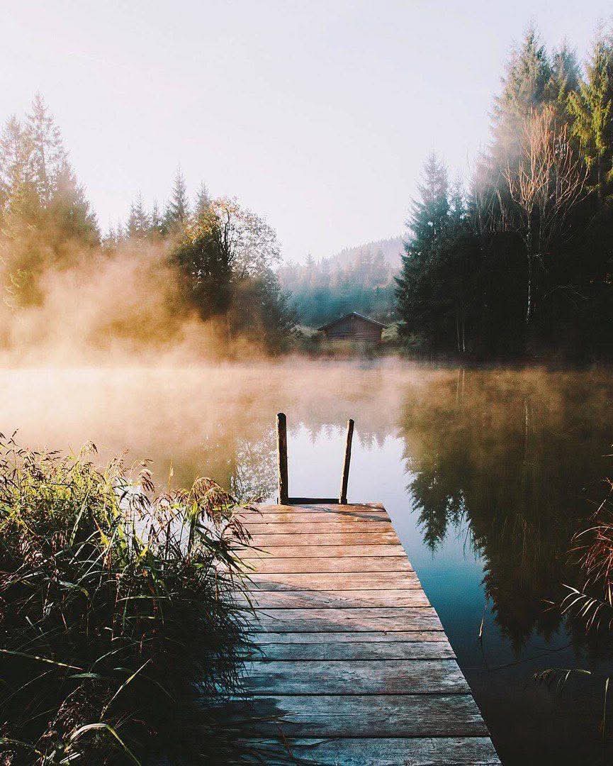 Wooden dock leading into a misty lake at sunrise with forested hills and a cabin in the background