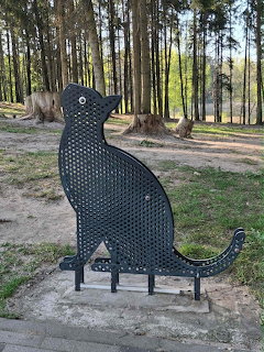 Silhouette of a sitting cat sculpture made from perforated black metal, positioned outdoors among trees