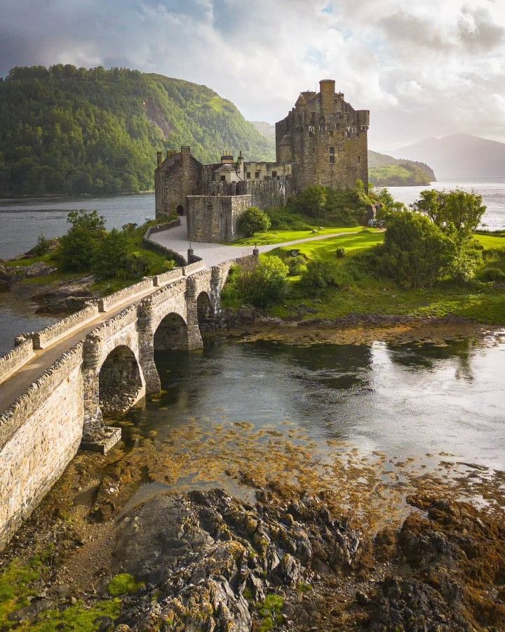 Medieval Scottish castle on island with stone bridge over water, green hills and dramatic sky