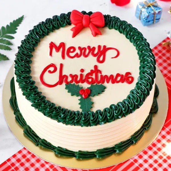 Round Christmas cake with white frosting, green wreath border, red bow on top, and Merry Christmas written in red icing