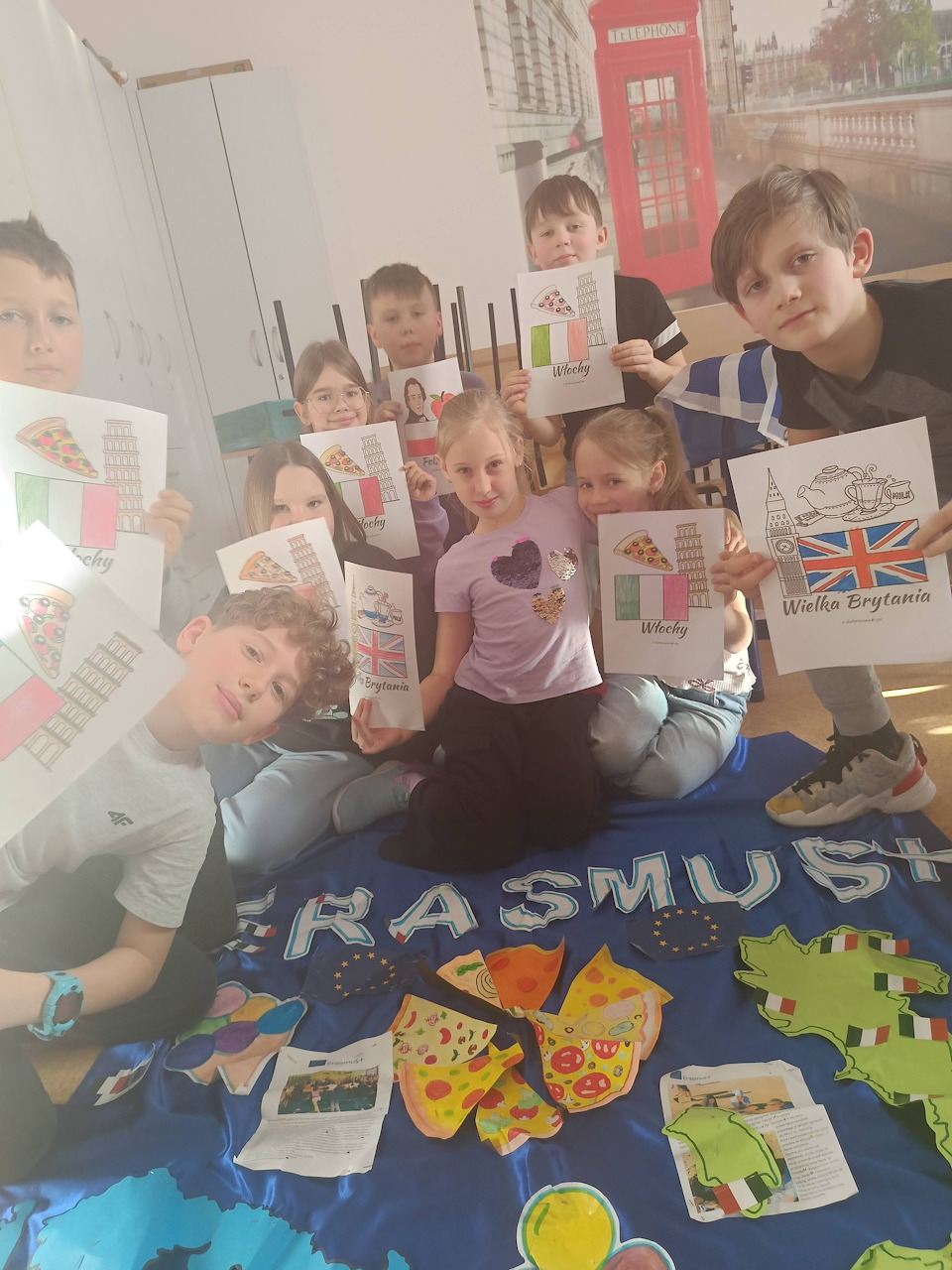 Group of children holding up their drawings in a classroom with an Erasmus banner on the table