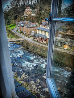Elevated window view of Lynmouth village with a rushing river, stone buildings, and woded hillside
