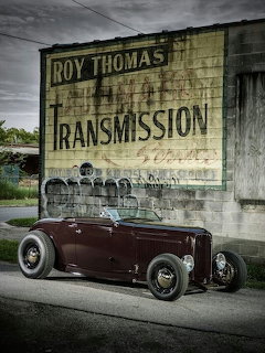 Dark red vintage hot rod roadster parked on street in front of old brick building with painted sign