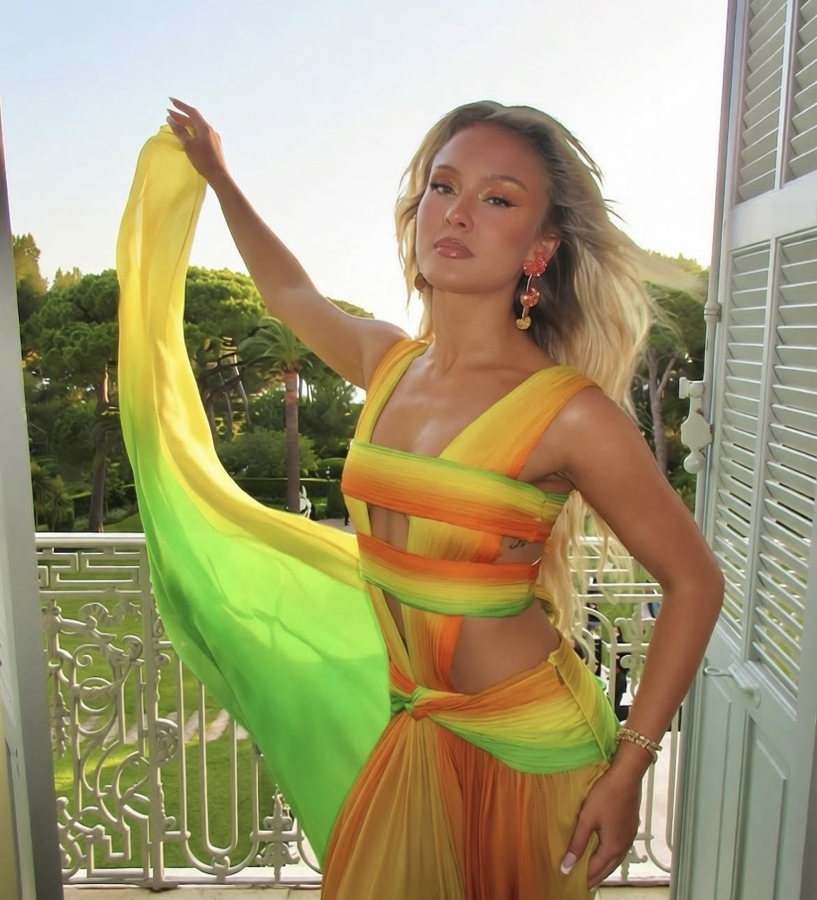Woman in yellow, orange and green ombre dress holding flowing fabric on white balcony