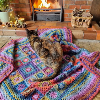 Tortoiseshell cat sitting on a colorful crocheted blanket by a brick fireplace