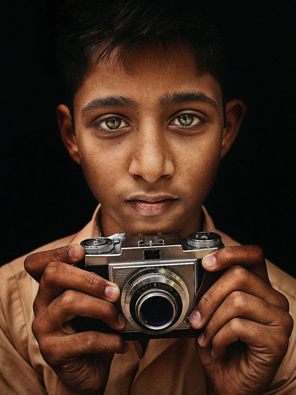 Young boy with green eyes holding a vintage film camera, close-up portrait on dark background