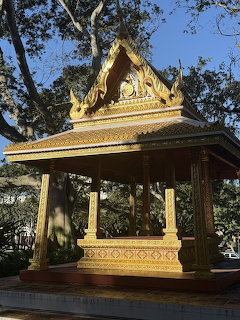 Golden Buddhist pagoda with ornate tiered roof and pillars in a tree-shaded courtyard
