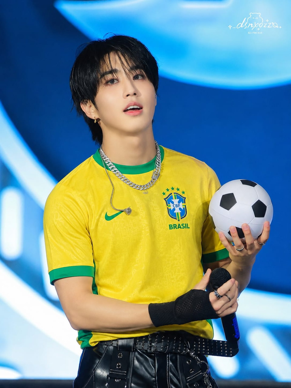 Young man in a yellow Brazil soccer jersey holding a soccer ball and microphone on stage