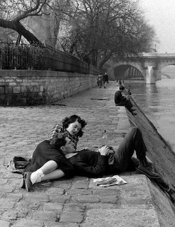 Black and white photo of a couple lounging on a cobblestone quay beside a river in Paris with a bridge behind