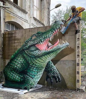3D street art of a giant crocodile with open mouth painted on a building wall with a person climbing above it