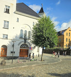 White church with pointed tower on cobblestone plaza with tree and colorful buildings in Bergen
