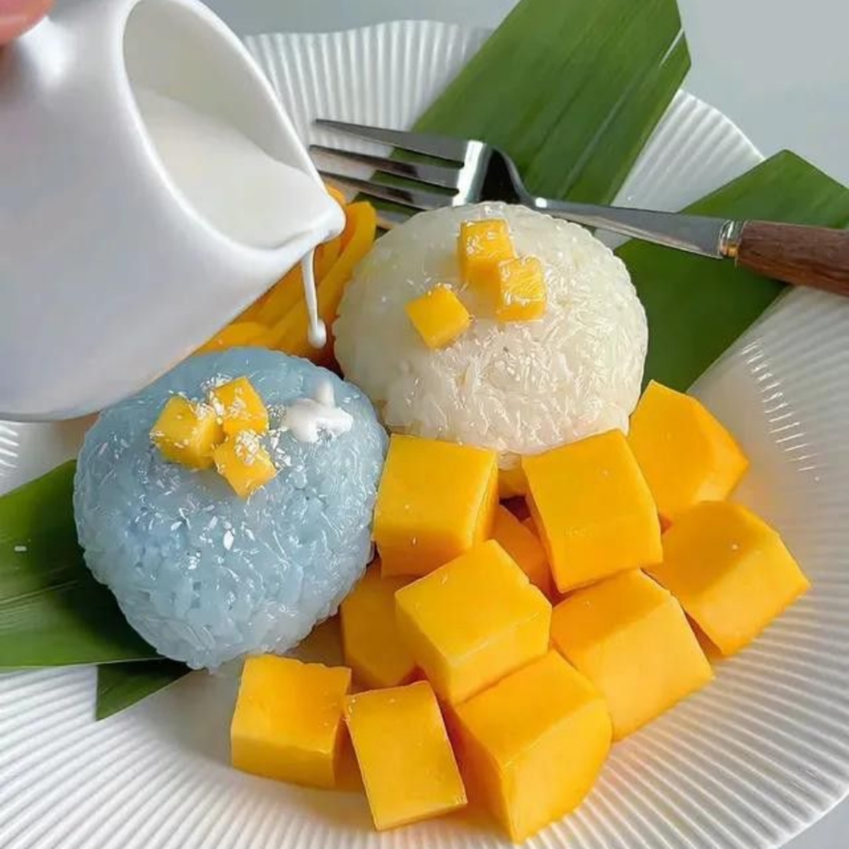 Mango sticky rice dessert with blue and white rice mounds, mango cubes, and coconut cream on banana leaf