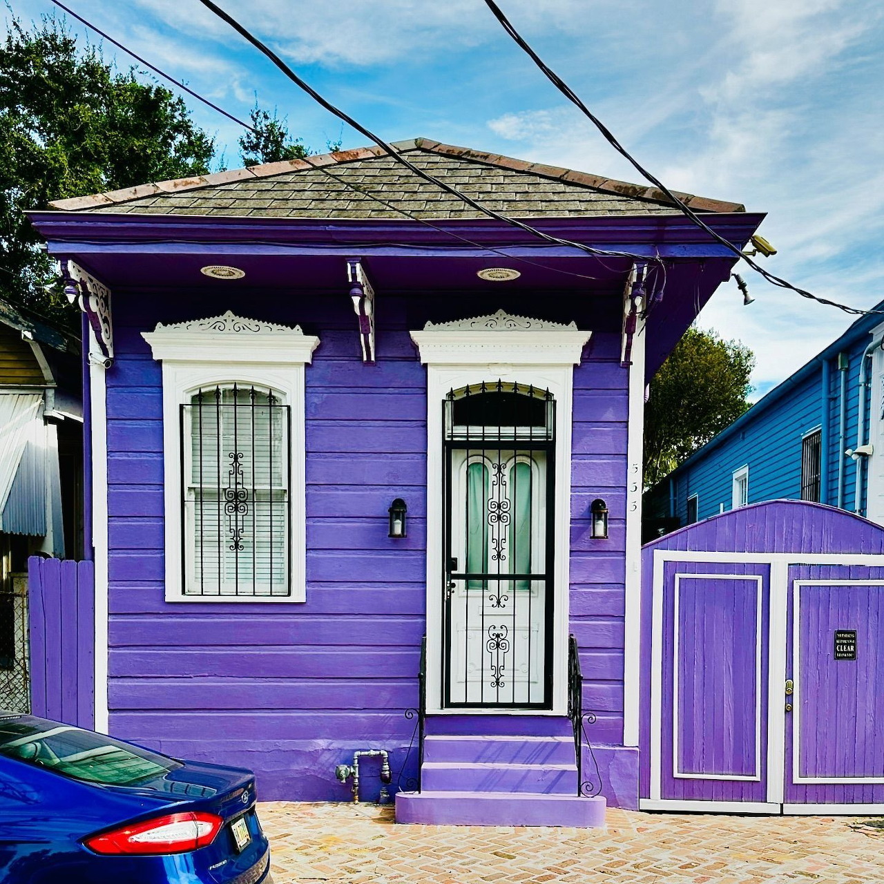 Small purple wooden house with white arched door and window, metal roof, and purple picket fence