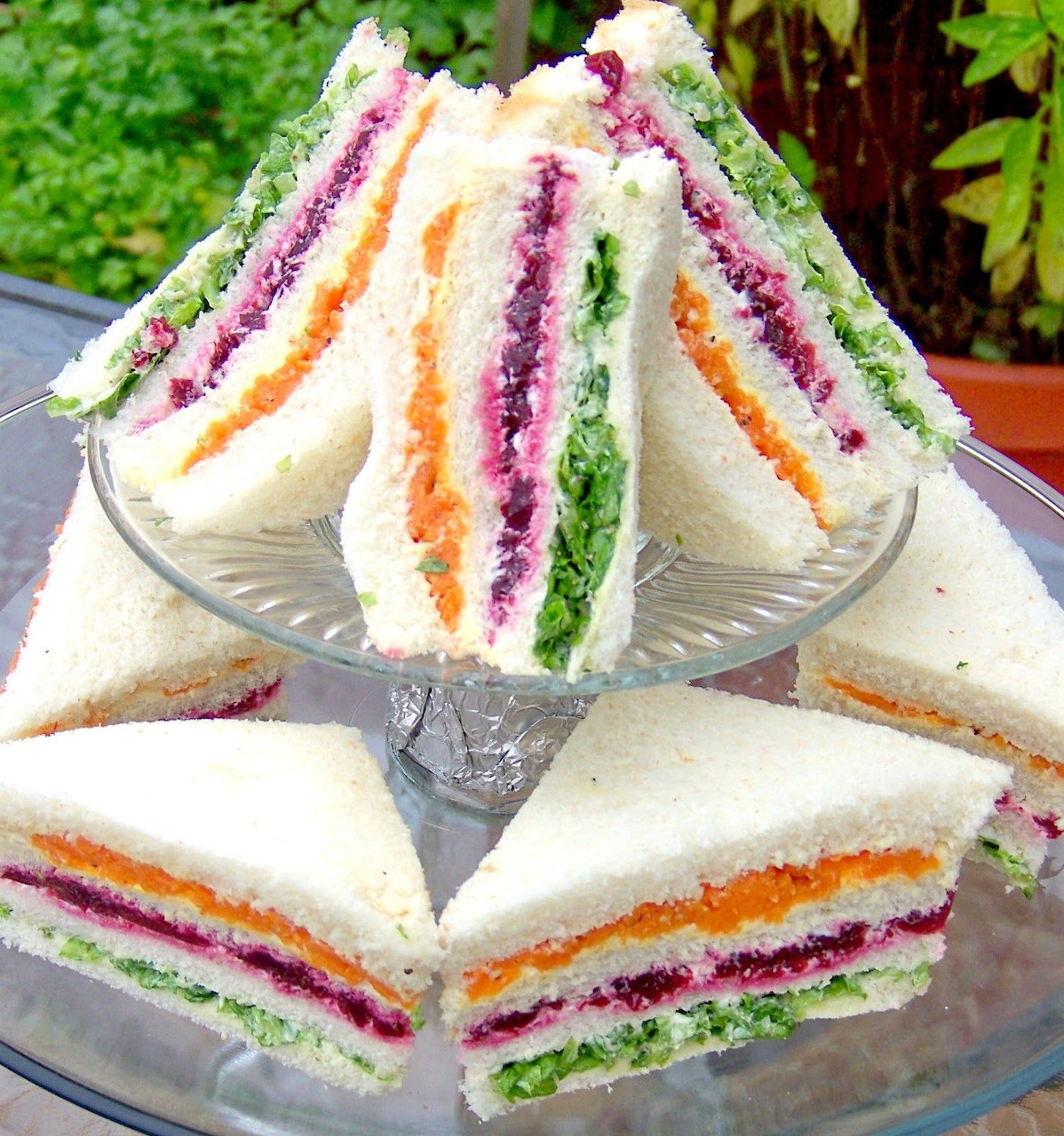 Layered ribbon sandwiches with colorful fillings stacked on glass cake stands outdoors