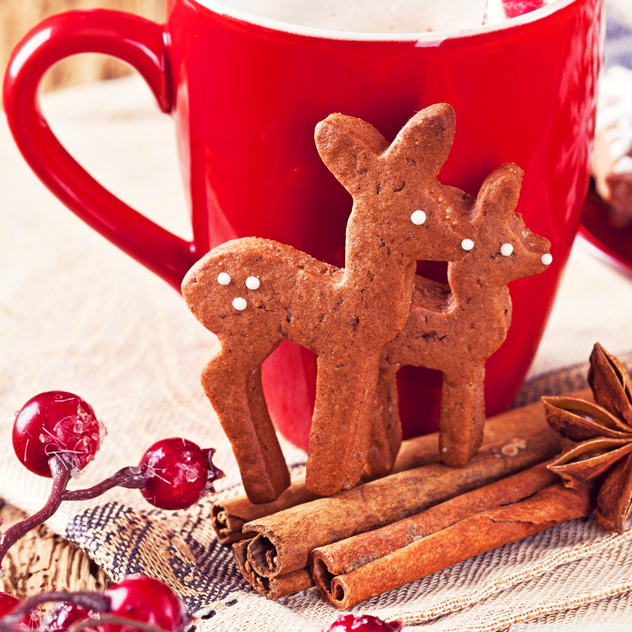 Gingerbread deer-shaped cookies with white icing dots next to red mug, cinnamon sticks, and holiday decorations