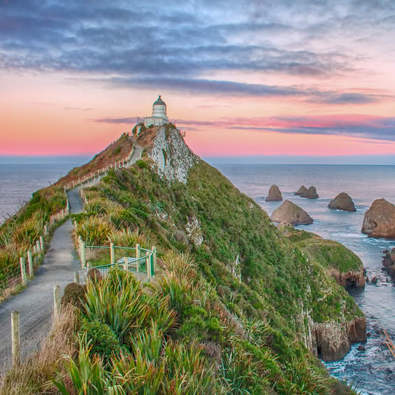 Lighthouse atop a green coastal cliff at sunset with a winding path and rocky sea stacks in the background.