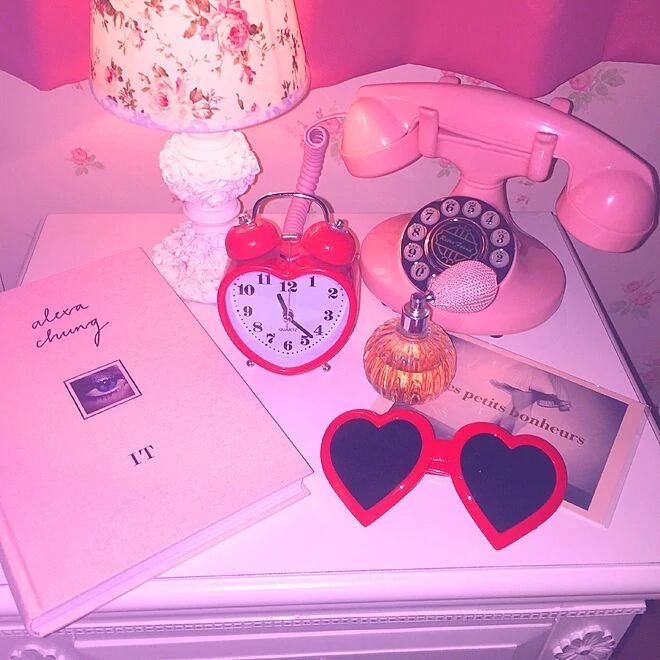 Pink tabletop still life with a retro phone, heart alarm clock, lamp, perfume, book, and heart sunglasses