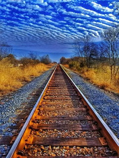 Railroad tracks extending toward horizon under blue sky with clouds, bordered by dry grass and bare trees